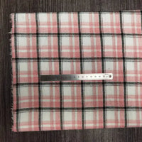 Une règle de 20 cm est posée horizontalement sur le tissu en flanelle à carreaux colorés au mètre de Longon Craft France, un tissu ultra-douillet en polyester-coton pour l'automne, présenté en rose, blanc et noir sur une surface brune texturée.