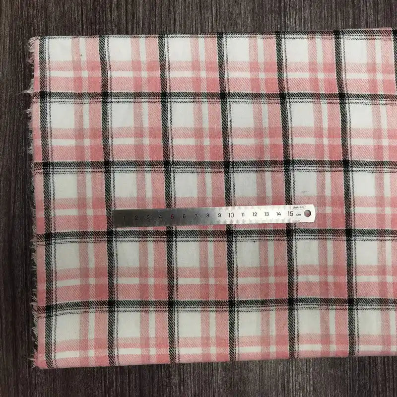 Une règle de 20 cm est posée horizontalement sur le tissu en flanelle à carreaux colorés au mètre de Longon Craft France, un tissu ultra-douillet en polyester-coton pour l'automne, présenté en rose, blanc et noir sur une surface brune texturée.