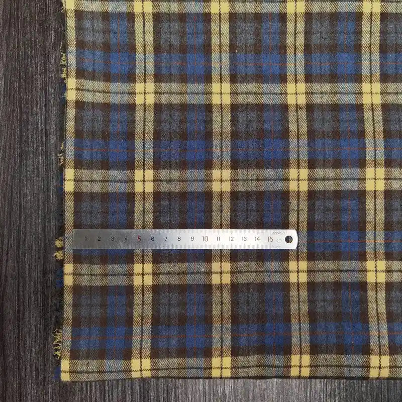 Un tissu en flanelle à carreaux colorés de Longon Craft France, idéal pour l'automne, est posé sur une surface en bois avec une règle métallique de 15 cm placée horizontalement le long du bord.