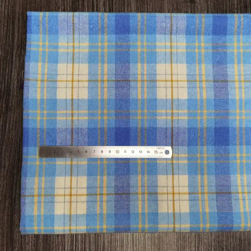 Un morceau plié de la flanelle à carreaux colorés au mètre de Longon Craft France, un tissu douillet en polyester-coton pour l'automne, repose sur du bois, surmonté d'une règle métallique de 15 cm.