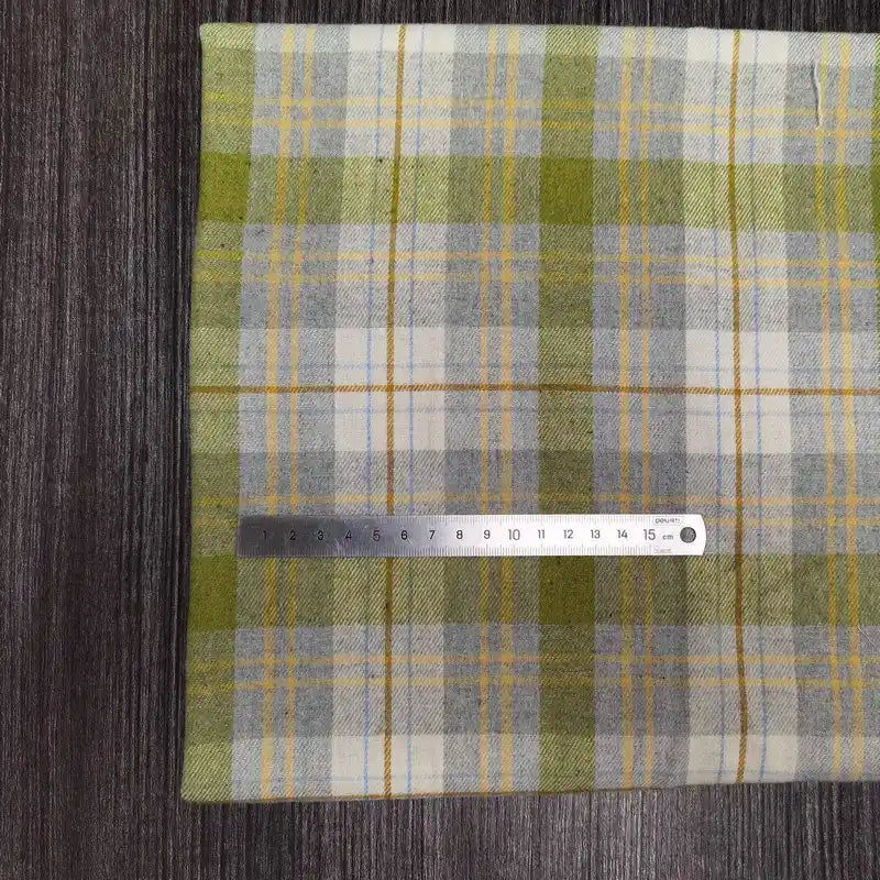 Une règle métallique de 15 cm repose sur un tissu en flanelle à carreaux verts, gris et blancs (Tissu en flanelle à carreaux colorés au mètre de Longon Craft France), en polyester-coton doux, présenté sur une surface grise texturée.