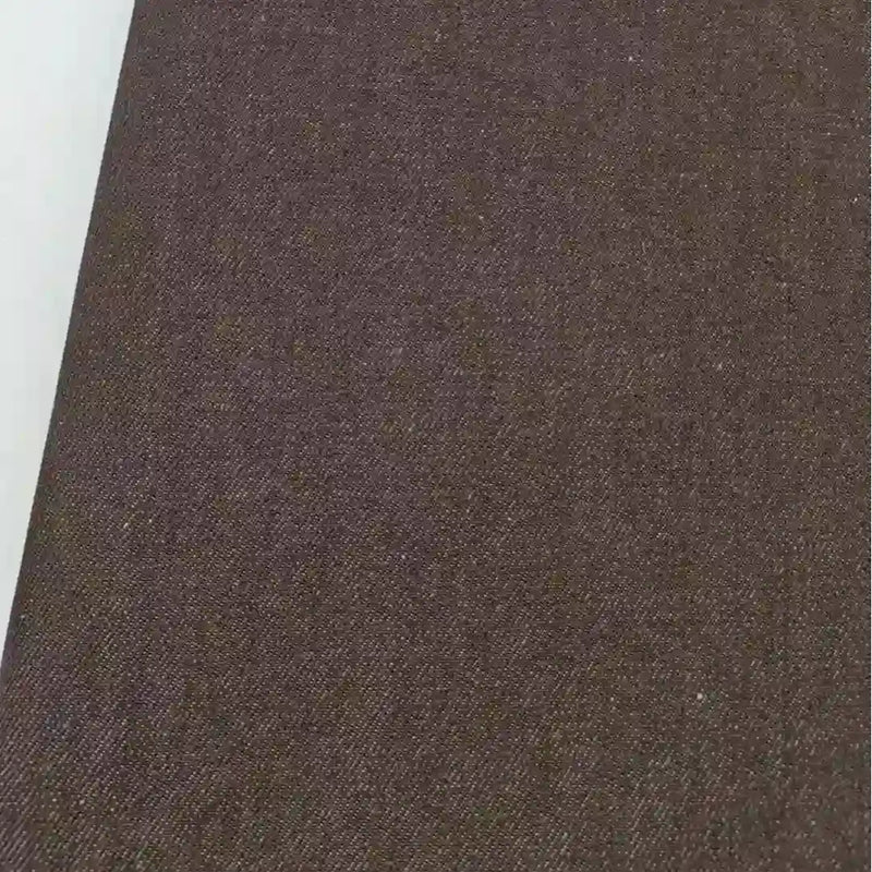 Gros plan sur le Tissu jean tissé coloré au mètre de Longan Craft France, un denim de coton de qualité supérieure de 6,5 oz sergé pour les jeans, montrant sa surface texturée brune avec un bord blanc sur la gauche.