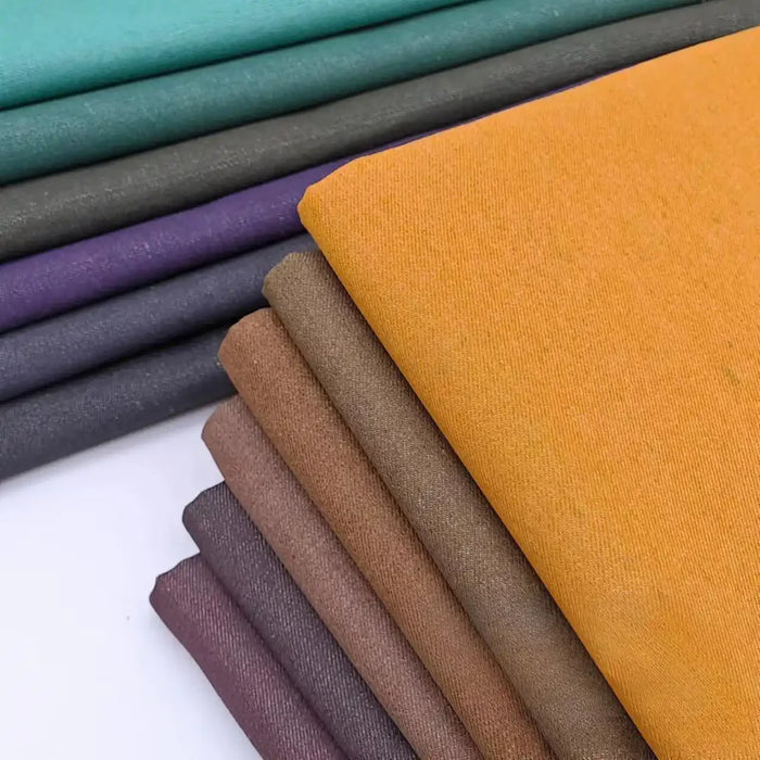 Une pile d'échantillons de denim colorés (jaune moutarde, marron, gris, violet, sarcelle) du Tissu jean tissé coloré au mètre 6.5 oz coton de Longan Craft France, disposés sur une surface blanche pour une décoration intérieure élégante.