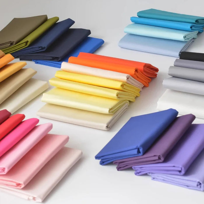 Des piles de popeline 100% coton pliée en rose, jaune, bleu, orange, violet, beige, gris et vert sont soigneusement disposées - idéales pour les chemises, les robes et les doublures. Popeline de coton au mètre par Longan Craft France.