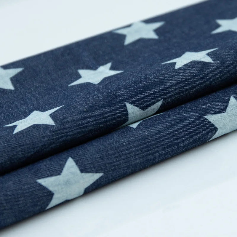 Tissu denim coton imprimé de fleurs et d'étoiles au mètre de Longan Craft France, en denim léger, présente un motif d'étoiles blanches sur fond bleu. Le tissu est soigneusement plié pour mettre en valeur le design répétitif.