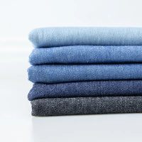 Une pile de cinq tissus denim délavés 6 oz au mètre de Longan Craft France, dans des tons bleus et gris, du plus clair au plus foncé, est disposée sur un fond blanc uni.
