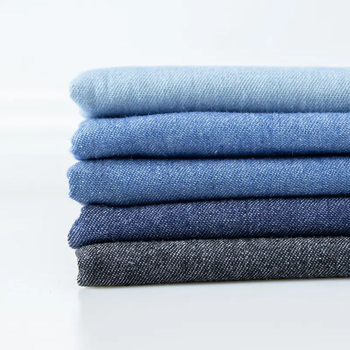 Une pile de cinq tissus denim délavés 6 oz au mètre de Longan Craft France, dans des tons bleus et gris, du plus clair au plus foncé, est disposée sur un fond blanc uni.