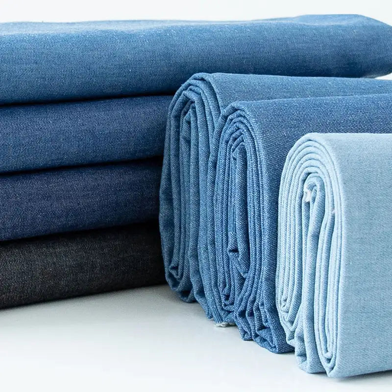 Plusieurs pièces de Longan Craft France Tissu denim délavé 6 oz au mètre (denim coton doux, tissu jean bleu) dans des tons bleus et gris sont disposées sur une surface blanche.