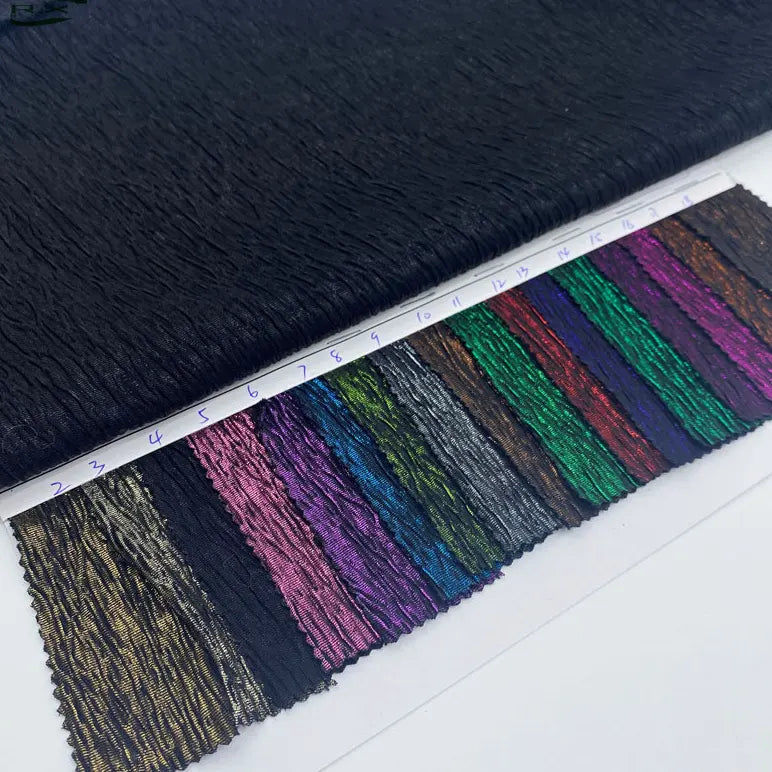 Un tissu lamé noir froissé au mètre de Longan Craft France est présenté au-dessus d'échantillons de tissus numérotés en or, bleu, rose, vert, rouge et violet - idéal pour les costumes, les robes et la couture.