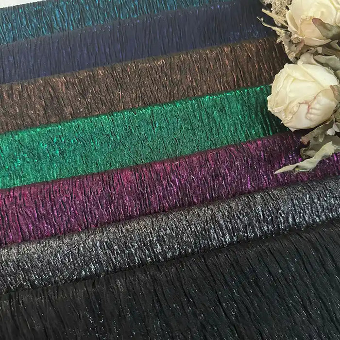 De multiples bandes de tissu lamé froissé de Longan Craft France en bleu, marron, vert et argent métallique sont disposées en rangées horizontales et rehaussées de roses artificielles de couleur crème dans le coin supérieur droit.