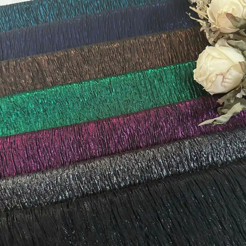 De multiples bandes de tissu lamé froissé de Longan Craft France en bleu, marron, vert et argent métallique sont disposées en rangées horizontales et rehaussées de roses artificielles de couleur crème dans le coin supérieur droit.