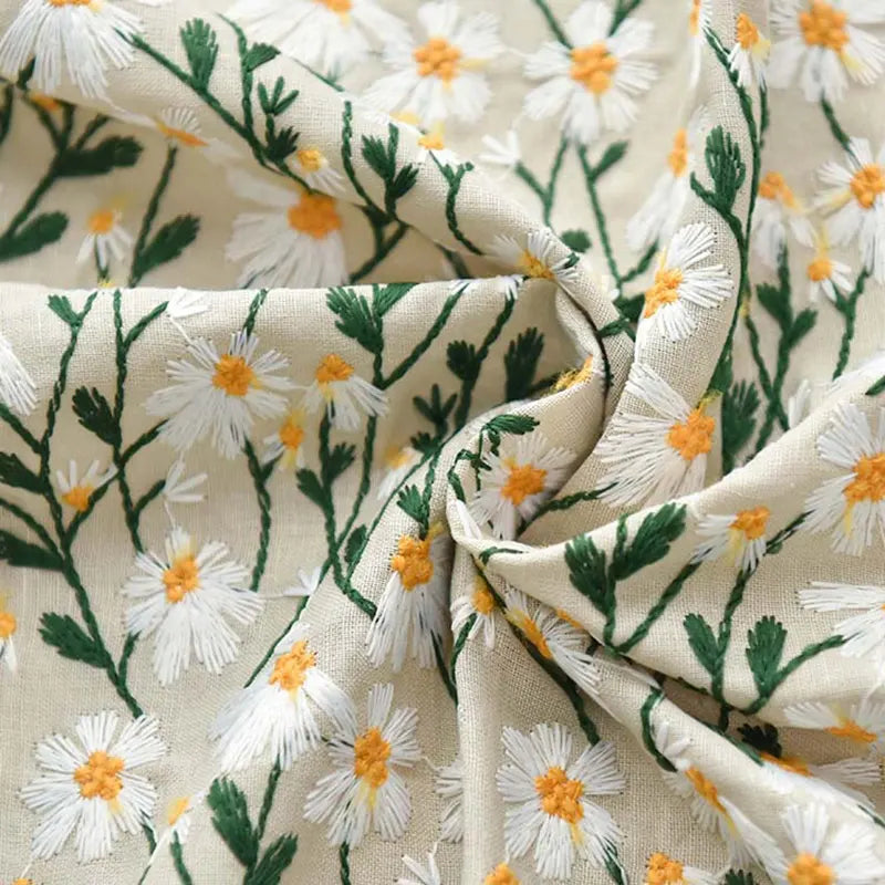 Un gros plan du tissu ramie marguerite de Longan Craft France montre une ramie de coton beige brodée de marguerites blanches et jaunes, de tiges vertes et de plis doux. Parfait pour les peignoirs, les rideaux, les projets créatifs et les tissus d'ameublement.