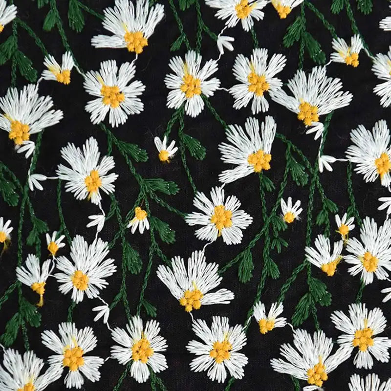 Gros plan sur le "Tissu ramie marguerite au mètre" de Longan Craft France, qui montre des fleurs brodées blanches et jaunes avec des tiges vertes sur un tissu doux en coton-ramie noir, idéal pour les robes, les rideaux ou les projets de couture créatifs.