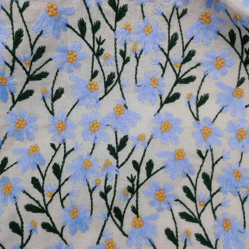 Le tissu ramie marguerite au mètre de Longan Craft France présente de délicates fleurs bleues brodées avec cœurs jaunes et tiges vertes sur fond beige, en fibres naturelles. Idéal pour robes, rideaux et loisirs créatifs.