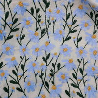 Le tissu ramie marguerite au mètre de Longan Craft France présente de délicates fleurs bleues brodées avec cœurs jaunes et tiges vertes sur fond beige, en fibres naturelles. Idéal pour robes, rideaux et loisirs créatifs.