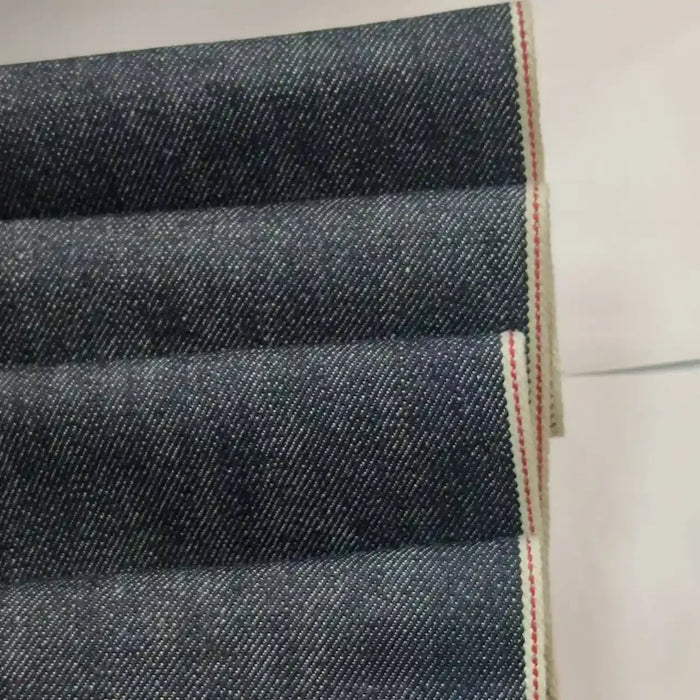 Le Tissu en jean selvedge bleu profond 14 oz au mètre de Longan Craft France, 100 % coton, présente une lisière blanche avec un liseré rouge. Parfait pour jeans, ce tissu haut de gamme est exposé plié sur une surface blanche.