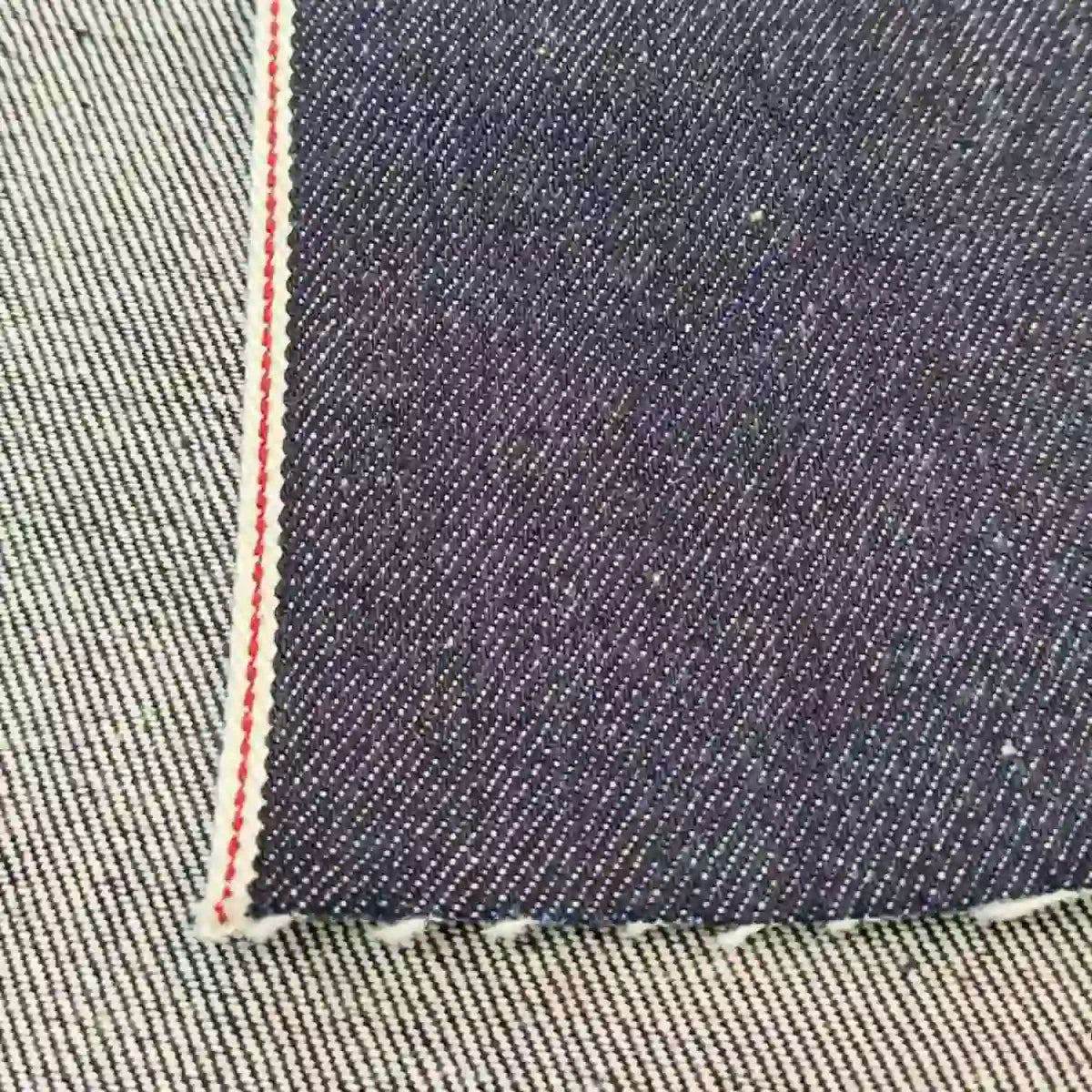 Gros plan sur le tissu en jean selvedge bleu profond 14 oz, 100% coton, de Longan Craft France, montrant son bord blanc et rouge sur un fond rayé bleu clair - une matière haut de gamme idéale pour les jeans.