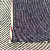 Gros plan sur le tissu en jean selvedge bleu profond 14 oz, 100% coton, de Longan Craft France, montrant son bord blanc et rouge sur un fond rayé bleu clair - une matière haut de gamme idéale pour les jeans.
