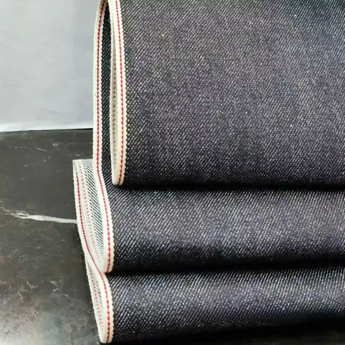 Trois pièces pliées du tissu en jean selvedge bleu profond 14 oz, 100 % coton, de Longan Craft France, présentent des bords blancs avec une ligne rouge, empilées sur une surface sombre sur un fond uni.