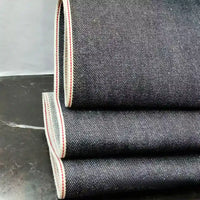 Trois pièces pliées du tissu en jean selvedge bleu profond 14 oz, 100 % coton, de Longan Craft France, présentent des bords blancs avec une ligne rouge, empilées sur une surface sombre sur un fond uni.