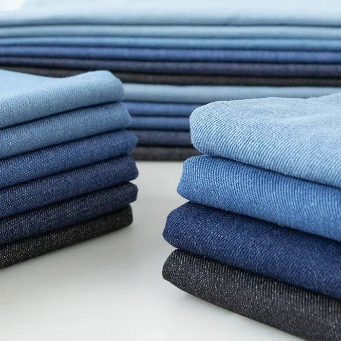 Des piles de tissu en jean extensible au mètre de Longan Craft France, y compris du denim stretch 8 oz pour vêtements et jeans, sont disposées proprement dans différentes nuances de bleu et de noir sur une surface blanche.