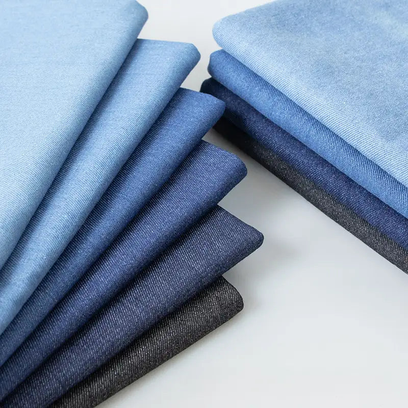 Des piles bien empilées de Tissu en jean extensible au mètre (8 oz) de Longan Craft France présentent différentes nuances de bleu et de gris, parfaites pour la fabrication de jeans et de vêtements.