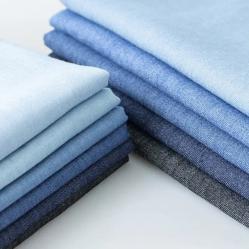 Une pile soigneusement empilée de tissu denim extensible 8 oz de Longan Craft France dans des tons allant du bleu clair au bleu marine, avec le "tissu en jean extensible au mètre", est disposée à plat et prête à être utilisée pour des projets de vêtements ou de jeans.
