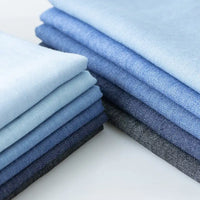 Une pile soigneusement empilée de tissu denim extensible 8 oz de Longan Craft France dans des tons allant du bleu clair au bleu marine, avec le "tissu en jean extensible au mètre", est disposée à plat et prête à être utilisée pour des projets de vêtements ou de jeans.