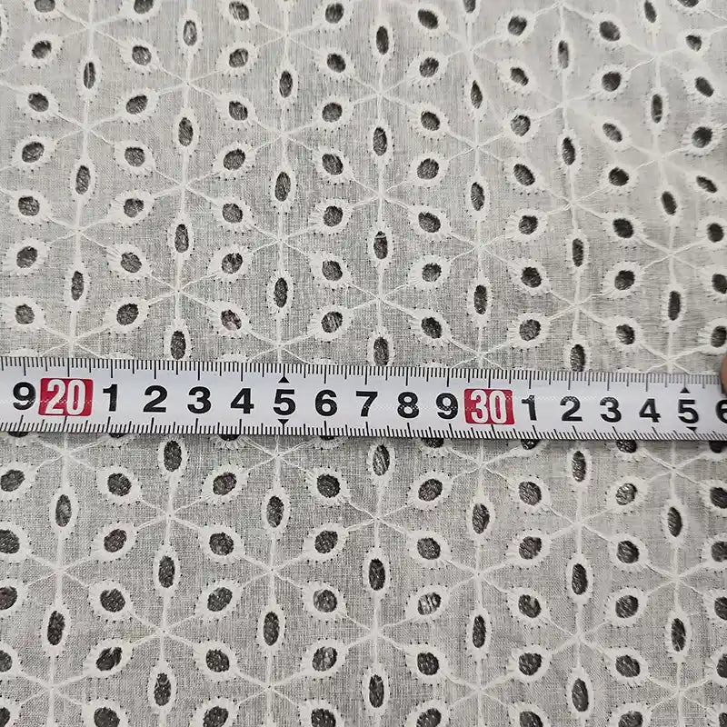Un mètre ruban en centimètres (20-34) est posé sur le voile de coton brodé au mètre de Longan Craft France, un tissu blanc avec une broderie florale ajourée, idéal pour les peignoirs, la couture et la décoration d'intérieur.