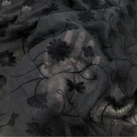 Une main est visible sous le Tissu brodé floral en gaze de lin au mètre de Longan Craft France. Ce tissu de lin naturel et transparent est délicatement brodé de fleurs et de lianes noires pour un effet semi-transparent.