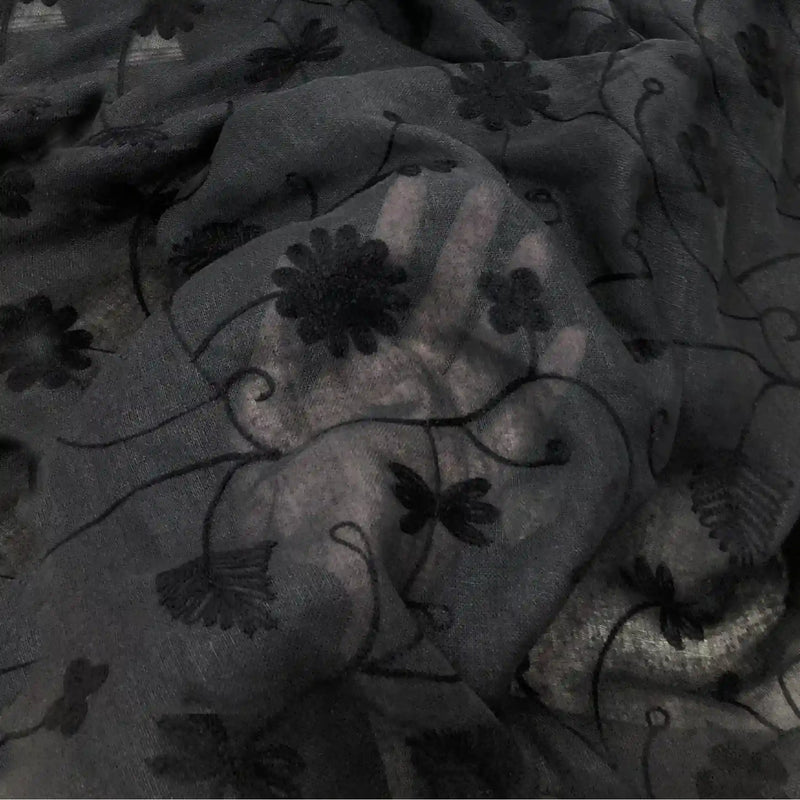 Une main est visible sous le Tissu brodé floral en gaze de lin au mètre de Longan Craft France. Ce tissu de lin naturel et transparent est délicatement brodé de fleurs et de lianes noires pour un effet semi-transparent.