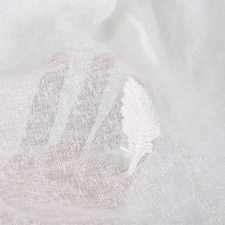 Une main sous le tissu brodé floral en gaze de lin au mètre de Longan Craft France révèle sa texture de lin fine et transparente et sa broderie délicate en forme de feuille, parfaite pour les vêtements d'été naturels.