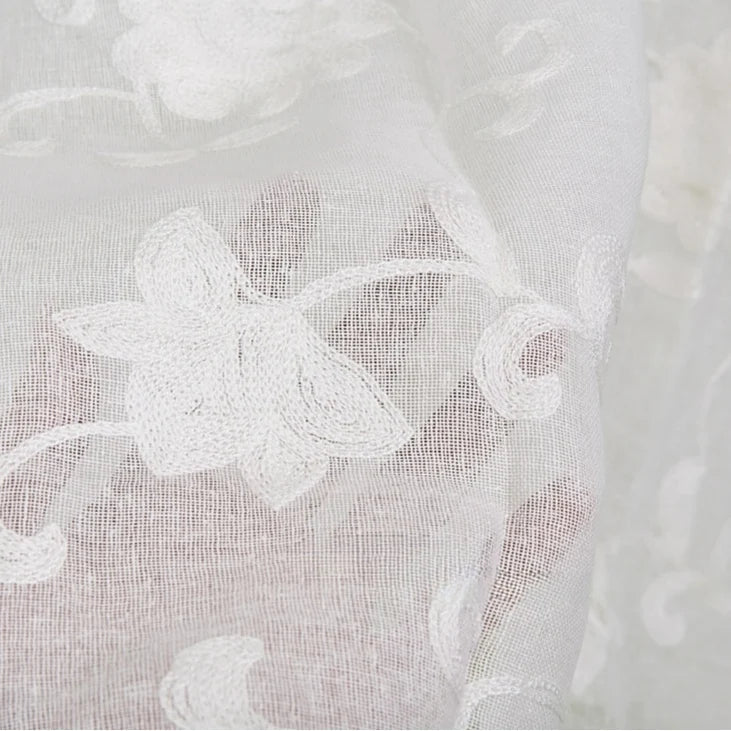Gros plan sur le Tissu brodé floral en gaze de lin au mètre de Longan Craft France, un tissu de lin naturel et transparent orné de fleurs et de vignes délicatement brodées en blanc. Le tissu transparent laisse transparaître l'arrière-plan.
