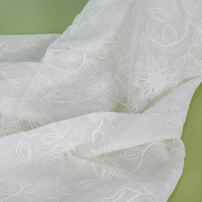 Gros plan sur le tissu brodé floral en gaze de lin au mètre de Longan Craft France, un tissu de lin naturel transparent aux motifs floraux et bouclés brodés, présenté sur un fond vert clair.