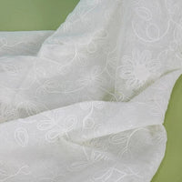 Gros plan sur le tissu brodé floral en gaze de lin au mètre de Longan Craft France, un tissu de lin naturel transparent aux motifs floraux et bouclés brodés, présenté sur un fond vert clair.