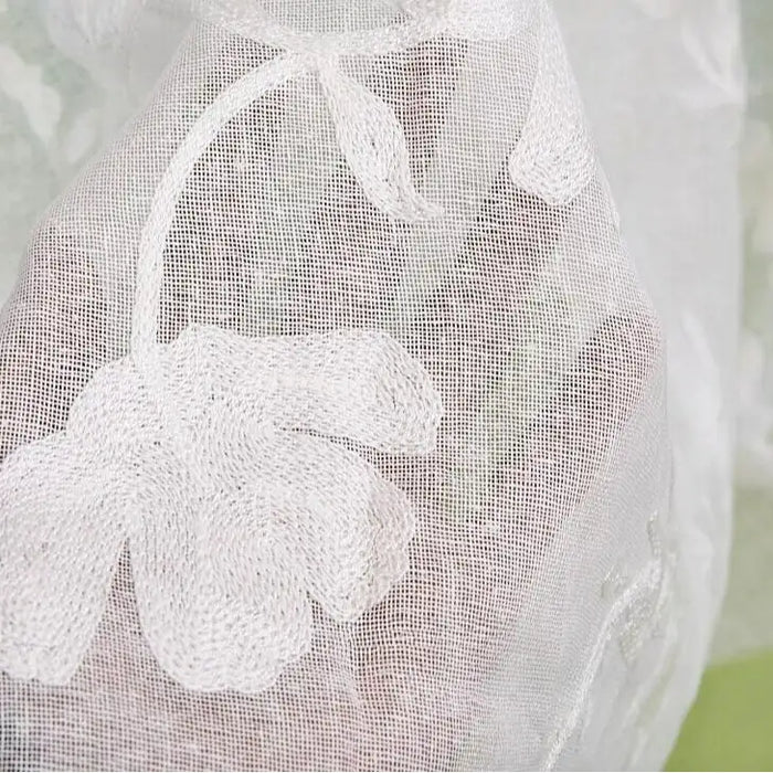 La main d'une personne est drapée de Tissu brodé floral en gaze de lin au mètre par Longan Craft France, un tissu de lin transparent et naturel avec un motif floral brodé, sur un fond doucement flou avec des touches de vert.