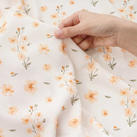 Une main tient délicatement le tissu mousseline à fleurs au mètre de Longan Craft France, un polyester imprimé de délicats motifs floraux dispersés orange clair et vert, idéal pour la création de vêtements.