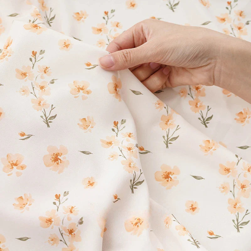 Une main tient délicatement le tissu mousseline à fleurs au mètre de Longan Craft France, un polyester imprimé de délicats motifs floraux dispersés orange clair et vert, idéal pour la création de vêtements.