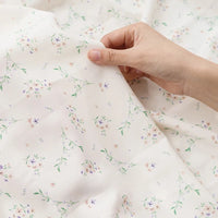 Une main tient Tissu mousseline à fleurs au mètre de Longan Craft France, un tissu blanc en polyester avec un délicat imprimé floral de feuilles vertes et de fleurs violettes, roses et jaunes, idéal pour la confection de vêtements.
