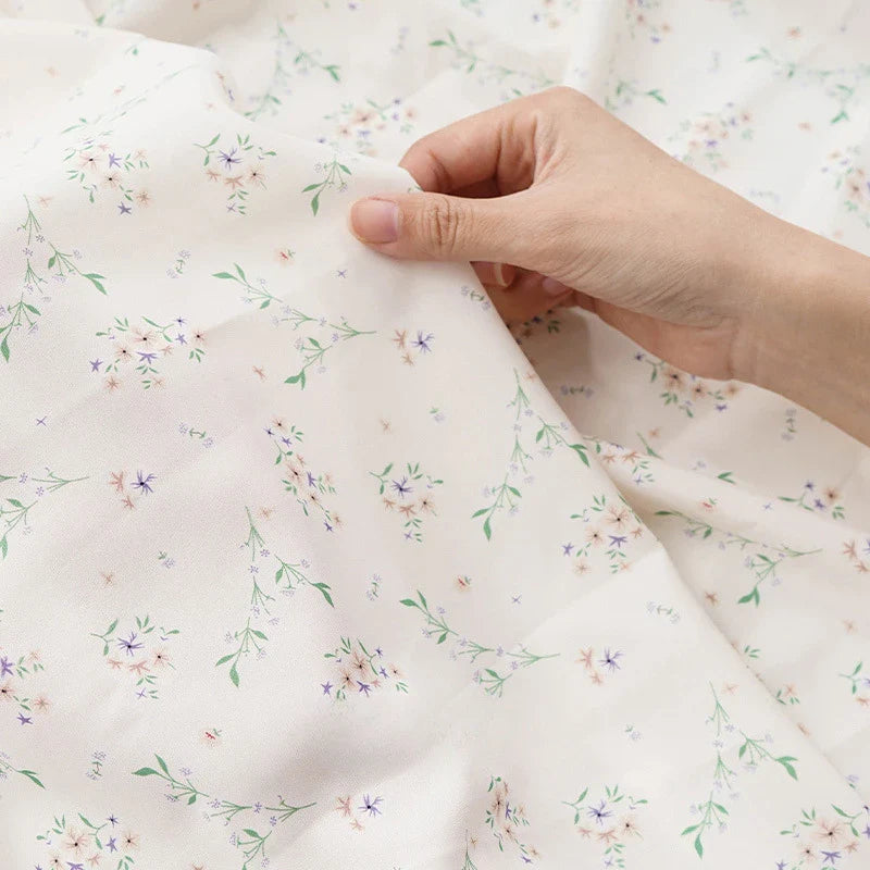 Une main tient Tissu mousseline à fleurs au mètre de Longan Craft France, un tissu blanc en polyester avec un délicat imprimé floral de feuilles vertes et de fleurs violettes, roses et jaunes, idéal pour la confection de vêtements.