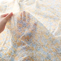 Une main présente le tissu mousseline à fleurs au mètre en polyester transparent et léger de Longan Craft France, avec un délicat imprimé floral jaune et bleu sur fond blanc, parfait pour la confection de vêtements.