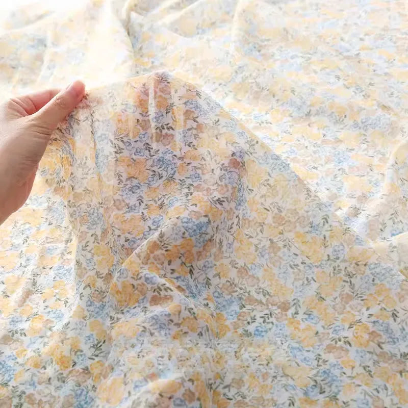 Une main présente le tissu mousseline à fleurs au mètre en polyester transparent et léger de Longan Craft France, avec un délicat imprimé floral jaune et bleu sur fond blanc, parfait pour la confection de vêtements.