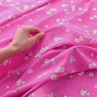 Une main tient délicatement Tissu mousseline à fleurs au mètre de Longan Craft France, un tissu polyester imprimé aux motifs petites marguerites blanches et fleurs bleues, idéal pour la confection de vêtements.