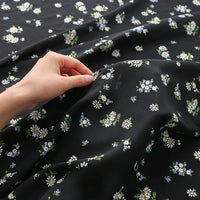 A hand holds Tissu mousseline à fleurs au mètre de Longan Craft France, en polyester imprimé avec un motif floral blanc, bleu et jaune, légèrement froissé et étalé sur une surface plane.