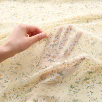 Une main brandit un morceau de Tissu mousseline à fleurs au mètre Longan Craft France, une mousseline de polyester jaune clair avec un imprimé floral blanc, jaune et vert. D'autres pièces de ce tissu aérien apparaissent à l'arrière-plan.