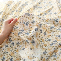 Une main tient délicatement le tissu mousseline à fleurs au mètre de Longan Craft France, un tissu léger en polyester avec des motifs floraux jaunes et bleus, idéal pour la confection de vêtements.