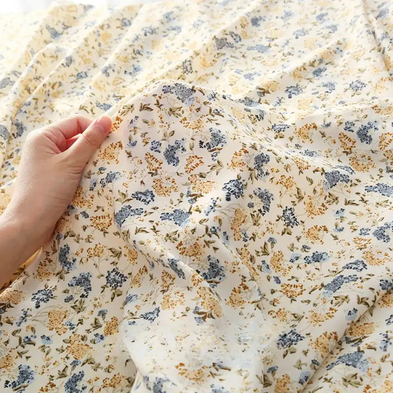 Une main tient délicatement le tissu mousseline à fleurs au mètre de Longan Craft France, un tissu léger en polyester avec des motifs floraux jaunes et bleus, idéal pour la confection de vêtements.