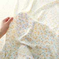 A hand displays a section of Tissu mousseline à fleurs au mètre de Longan Craft France, en polyester imprimé avec des petites marguerites jaunes, tandis qu'en arrière-plan, davantage de cette mousseline délicate est étalée à plat.