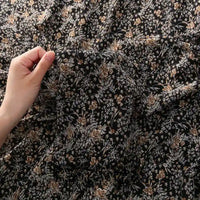 Une main tient le tissu mousseline à fleurs au mètre de Longan Craft France, un tissu léger en polyester noir avec un délicat imprimé de petites fleurs en marron, beige et blanc, parfait pour la confection de vêtements. La mousseline semble légèrement froncée.