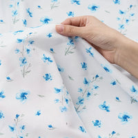 Une main tient le tissu mousseline à fleurs au mètre de Longan Craft France, un tissu polyester délicat avec de petites fleurs bleues et des feuilles gris verdâtre, parfait pour les vêtements.
